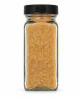 A clear glass spice jar filled with a golden-brown granular seasoning blend, sealed with a black ribbed screw-top lid, isolated on a white background.