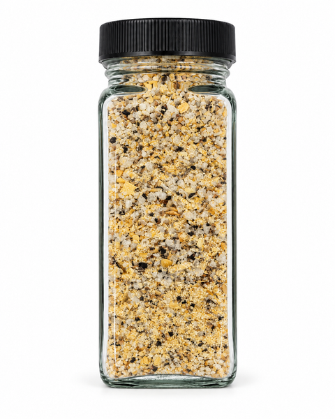 A clear glass spice jar filled with a coarse yellow and beige seasoning blend, topped with a black ribbed screw cap, isolated on a white background.