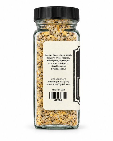 A clear glass spice jar with a black ribbed lid, showing the back of the label. The label is cream-colored with black text detailing usage suggestions for eggs, wings, steak, burgers, fries, veggies, pulled pork, asparagus, avocado, potatoes, the brand Street City Ball, address, website, and 'Made in USA'.