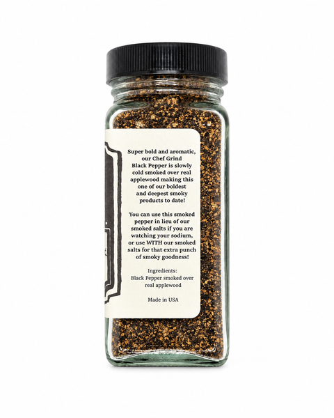 A clear glass spice jar with a black screw cap and a cream paper label, filled with black pepper smoked over real applewood. The label features descriptive text in black font.